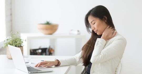Asian woman sitting at work desk, holding neck in pain